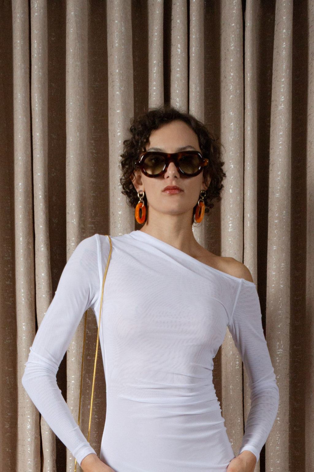 Woman wearing a white off-shoulder top with sunglasses and earrings against a beige curtain background