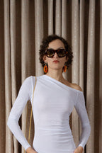 Woman wearing a white off-shoulder top with sunglasses and earrings against a beige curtain background