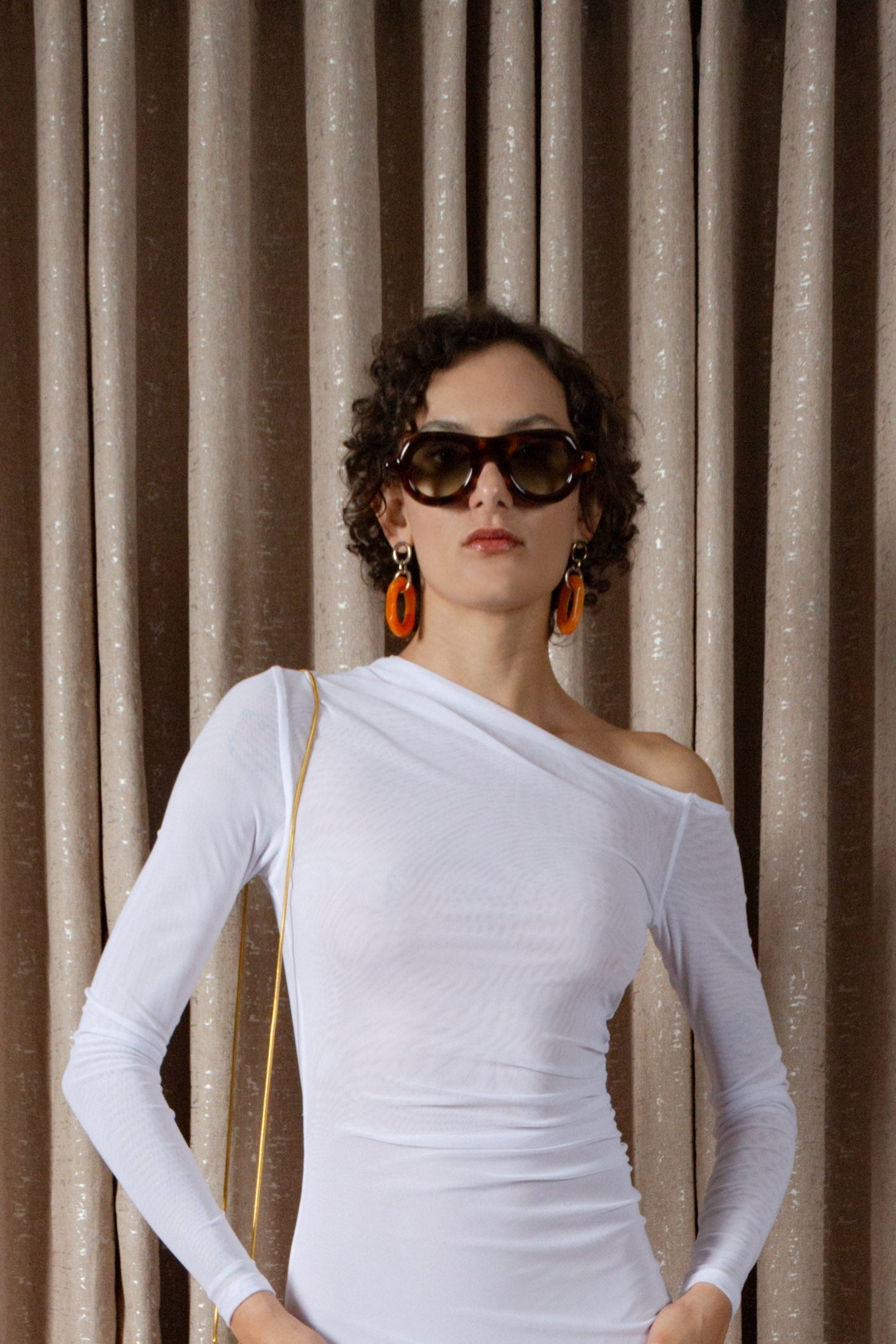 Woman wearing a white off-shoulder top with sunglasses and earrings against a beige curtain background